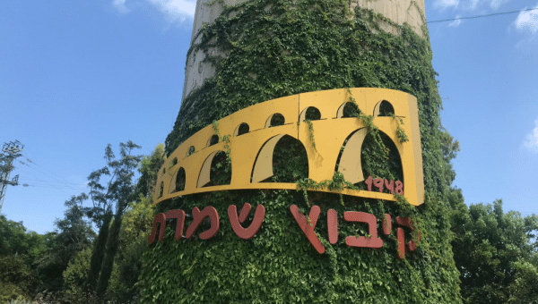 שער הכניסה הראשי לקיבוץ שמרת ביום בהיר, עם צמחייה מטופחת ושילוט היישוב.