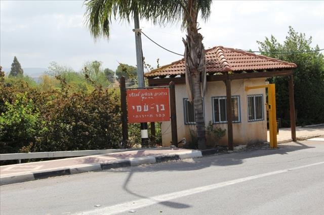 שלט הכניסה האדום של המושב "ברוכים הבאים לבן עמי", הניצב בפתח היישוב על רקע צמחייה מטופחת.
