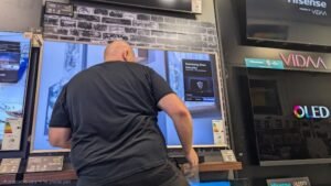 טכנאי מתקין מסך טלוויזיה גדול על קיר במשרד או בעסק, תוך הקפדה על חיבורים נקיים ותלייה בטיחותית ומאוזנת.